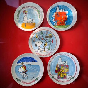 May include: Five vintage Christmas plates featuring Snoopy in various holiday scenes. Each plate has a different year: 1975, 1977, 1980, 1981, and 1982. The plates have a white border with a red trim and are set against a red background.