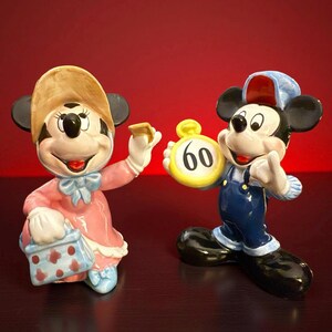 May include: Two ceramic figurines of cartoon characters. One is a female character in a pink dress and hat, holding a purse and a small object. The other is a male character in blue overalls and a cap, holding a yellow clock with the number 60.