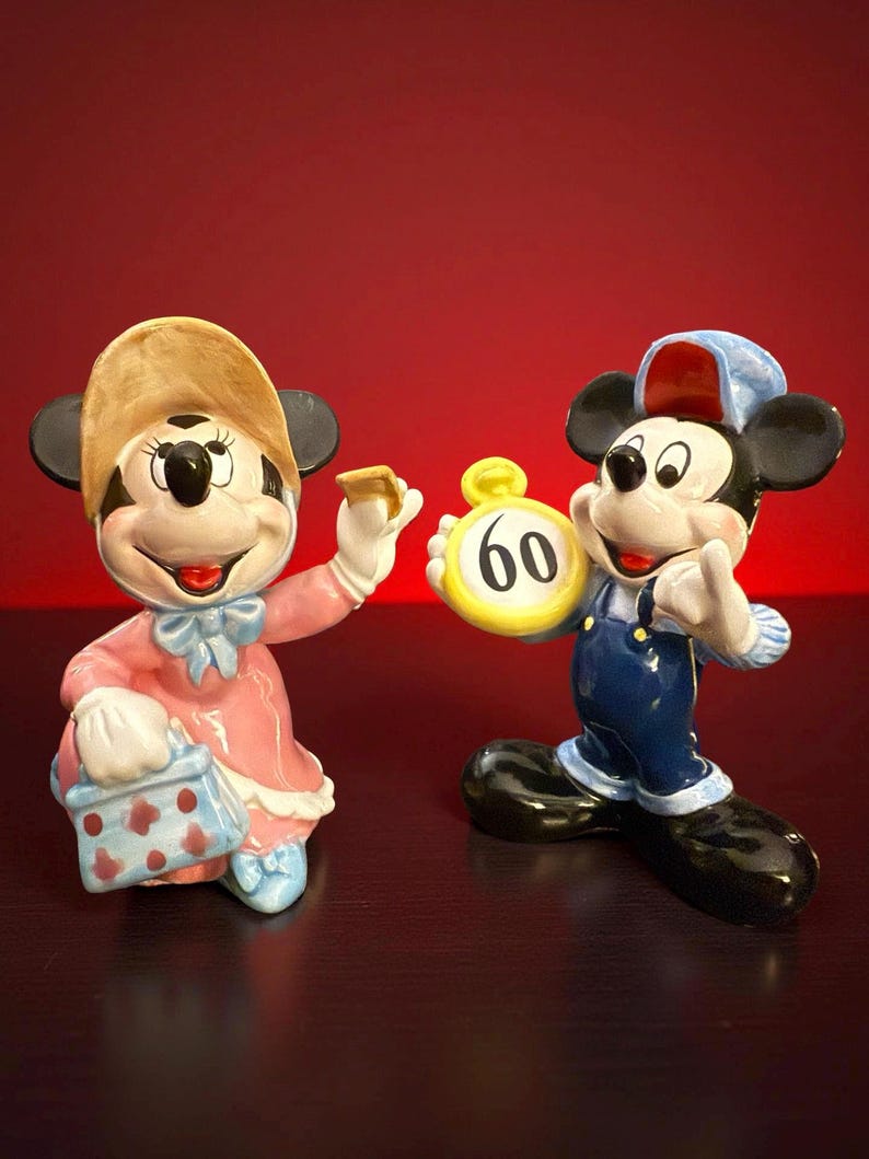 May include: Two ceramic figurines of cartoon characters. One is a female character in a pink dress and hat, holding a purse and a small object. The other is a male character in blue overalls and a cap, holding a yellow clock with the number 60.