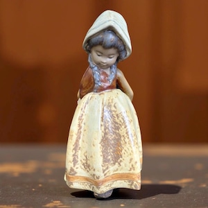 May include: A ceramic figurine of a girl wearing a bonnet and a long dress. The figurine has a light yellow dress with a brown trim, a brown top, and a white bonnet. The girl is looking down.