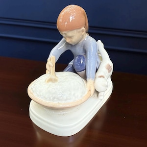 May include: A porcelain figurine depicting a child stirring a bowl, with a dog beside them. The child wears a blue outfit and a brown cap. The bowl is white with a light brown rim, and the base is white.