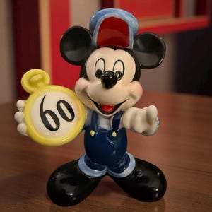 May include: A ceramic Mickey Mouse figurine wearing blue overalls and a conductor's hat, holding a yellow clock with the number 60. The figurine has black ears and shoes, and a smiling face.