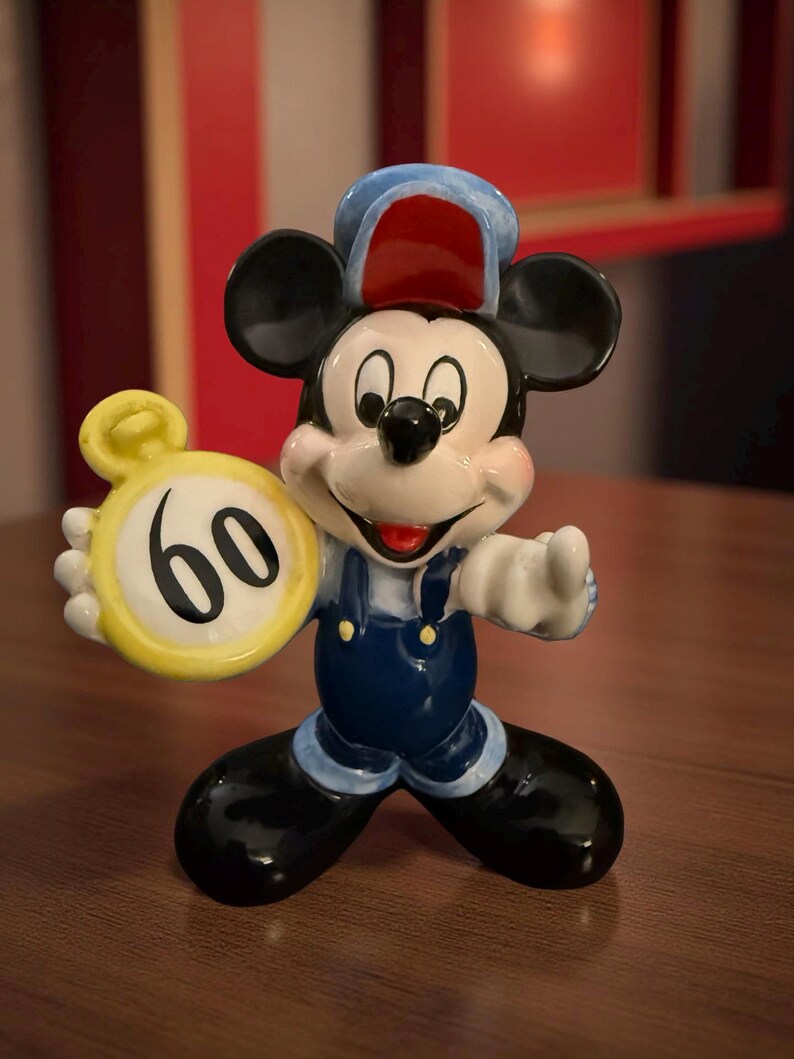 May include: A ceramic Mickey Mouse figurine wearing blue overalls and a conductor's hat, holding a yellow clock with the number 60. The figurine has black ears and shoes, and a smiling face.
