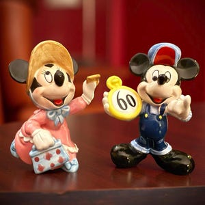 May include: Two ceramic figurines of Mickey and Minnie Mouse. Minnie is wearing a pink dress and a hat, holding a small purse. Mickey is in blue overalls and a hat, holding a yellow clock with the number 60.