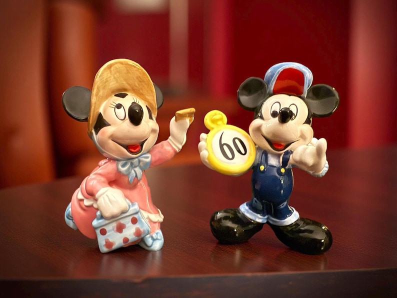 May include: Two ceramic figurines of Mickey and Minnie Mouse. Minnie is wearing a pink dress and a hat, holding a small purse. Mickey is in blue overalls and a hat, holding a yellow clock with the number 60.