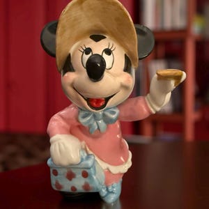 May include: A ceramic Minnie Mouse figurine, wearing a pink dress, blue bow, and a tan hat. The figurine is holding a small, round object in one hand and a blue and white polka-dotted basket in the other. The figurine has black ears and a smiling expression.