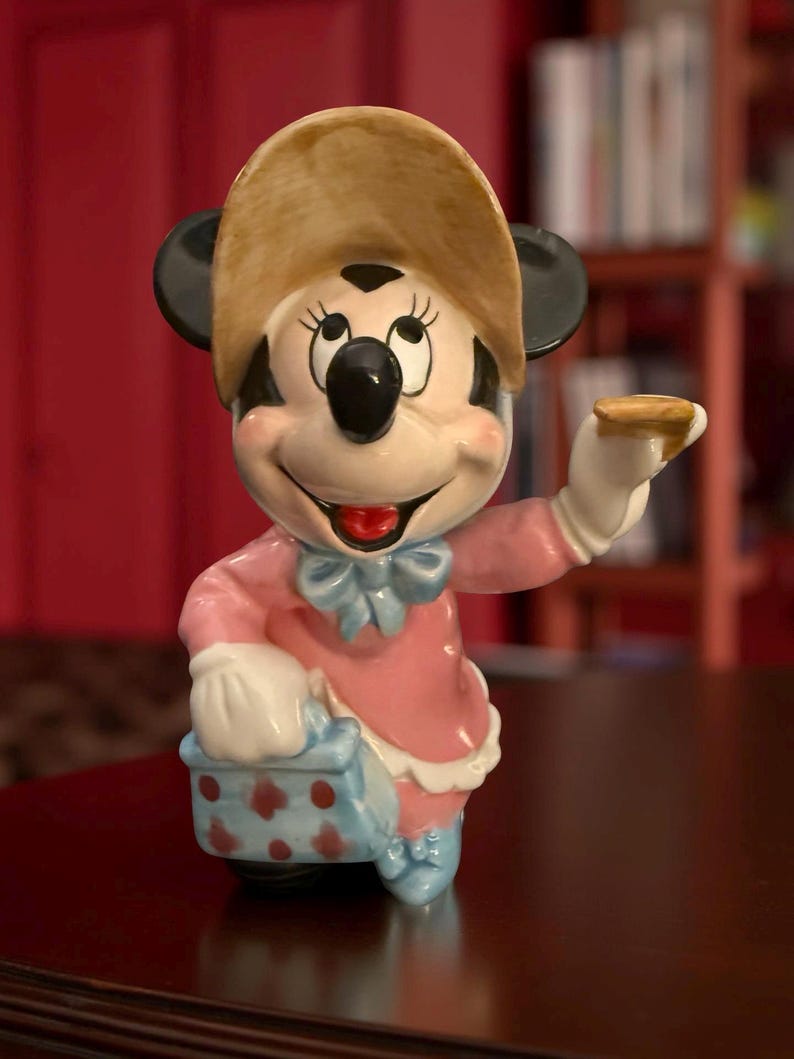 May include: A ceramic Minnie Mouse figurine, wearing a pink dress, blue bow, and a tan hat. The figurine is holding a small, round object in one hand and a blue and white polka-dotted basket in the other. The figurine has black ears and a smiling expression.