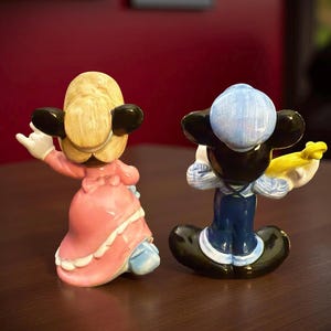 May include: Two ceramic figurines of cartoon characters. One is a female character in a pink dress with white trim, the other is a male character in a blue outfit. Both have black ears and are posed with their backs to the viewer.