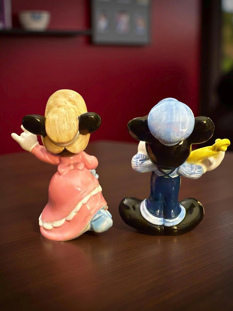 May include: Two ceramic figurines of cartoon characters. One is a female character in a pink dress with white trim, the other is a male character in a blue outfit. Both have black ears and are posed with their backs to the viewer.