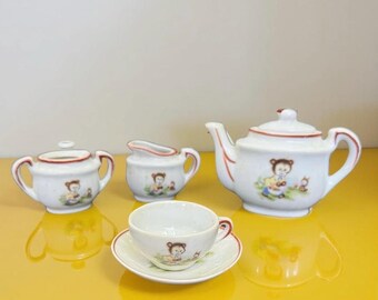 May include: A white ceramic tea set with a teapot, sugar bowl, creamer, cup, and saucer. Each piece features a brown bear illustration with red trim. The set is on a yellow surface.