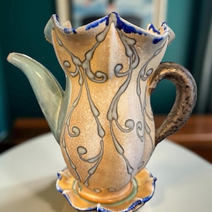 May include: A decorative ceramic teapot with a unique, stylized design. The teapot features a light tan body with blue and gray swirling patterns, a light green spout, and a dark brown handle. It sits on a matching saucer with blue and yellow accents.