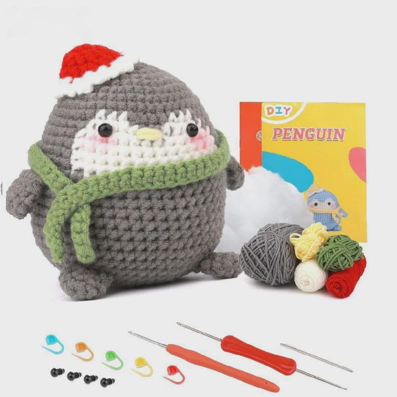 May include: A crocheted penguin wearing a red Santa hat and green scarf. The penguin is part of a DIY craft kit, with yarn, crochet hooks, and a booklet labeled "DIY PENGUIN". Includes stitch markers.