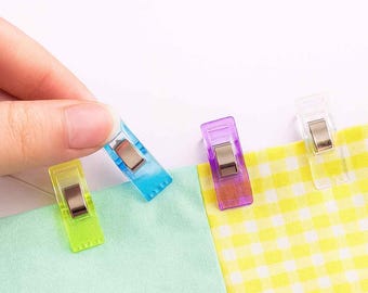 May include: A hand holding a blue sewing clip, with three other colorful clips on fabric. The clips are transparent and have metal clasps. The fabric is light green and yellow checkered.