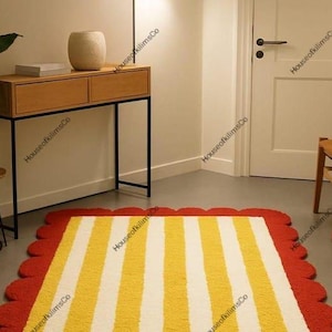 Peut inclure: Un tapis rectangulaire orné de rayures verticales jaunes et blanches avec une bordure festonnée rouge. Le tapis est posé sur un sol lisse dans une pièce comprenant une console en bois, une plante en pot et une chaise en bois.