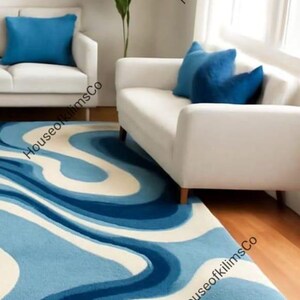 May include: A blue and white patterned rug with a wavy design, placed in a living room setting. The rug is the focal point, complemented by white sofas, blue cushions, and a green plant. The room has hardwood floors.