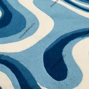 May include: A close-up of a rug with a wavy pattern in shades of blue and white. The rug has a soft, textured appearance. The design features flowing, organic shapes, creating a visually appealing and modern aesthetic.