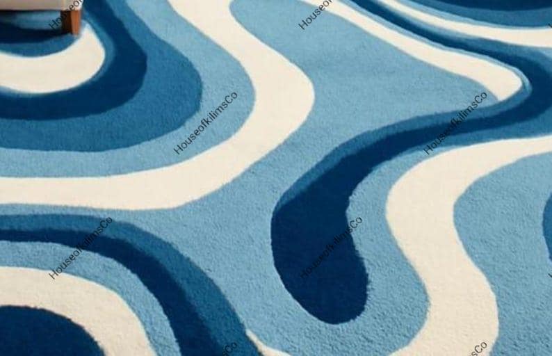 May include: A close-up of a rug with a wavy pattern in shades of blue and white. The rug has a soft, textured appearance. The design features flowing, organic shapes, creating a visually appealing and modern aesthetic.