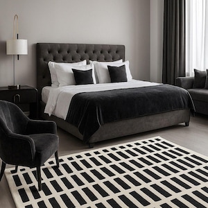 May include: A modern bedroom scene featuring a tufted headboard bed with white and black bedding. A black and white geometric patterned rug covers the floor. A black armchair and a side table with a lamp complete the room's decor.