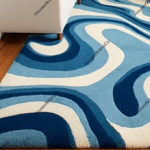 May include: A blue and white patterned rug with a wavy design. The rug features shades of blue and white, creating a flowing, abstract pattern. The rug is placed on a wooden floor, with a white sofa visible in the background.