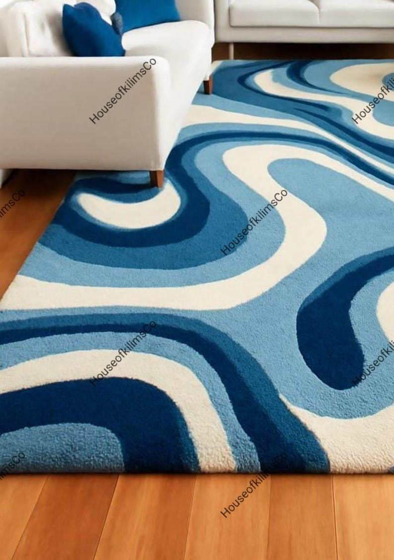 May include: A blue and white patterned rug with a wavy design. The rug features shades of blue and white, creating a flowing, abstract pattern. The rug is placed on a wooden floor, with a white sofa visible in the background.