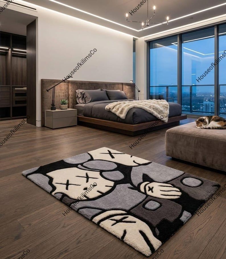 May include: A modern bedroom featuring a large bed with gray bedding and a plush throw. A gray and black cartoon character rug is on the wooden floor. A cat rests on a matching ottoman near a large window with a city view.