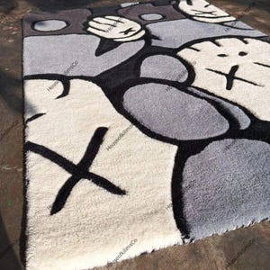 May include: A rectangular rug with a cartoon-style design in shades of gray, black, and off-white. The rug features a repeating pattern of abstract shapes and the letter "X". The rug is made of a soft, textured material.