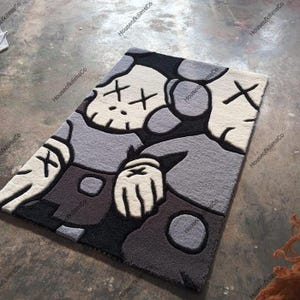 May include: A rectangular rug featuring a cartoon character design in shades of gray, black, and off-white. The rug has a plush texture and a repeating pattern of the character's face and body. The design includes cross-shaped eyes.