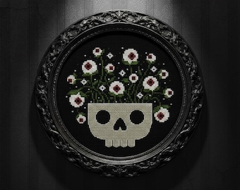 May include: A framed cross-stitch design featuring a skull-shaped vase filled with white flowers with red centers and green stems. The artwork is set against a black background and framed in a black, ornate oval frame.