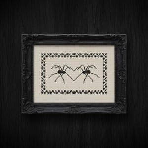 May include: A framed cross-stitch artwork featuring two spiders forming a heart shape. The spiders and heart are stitched in black thread against a cream-colored background. The frame is ornate and black, with a dark wood grain background.