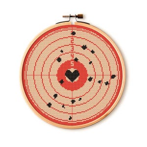 May include: A cross-stitch embroidery hoop featuring a target design with a heart in the center. The target has concentric circles in beige and red, with black bullet hole details. The wooden hoop is visible.