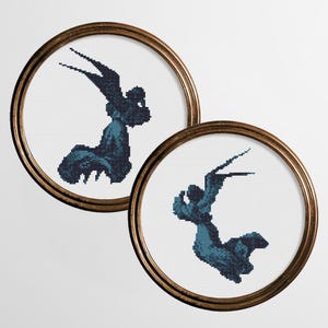 May include: Two framed cross-stitch artworks. Each features a dark teal figure with outstretched wings and a seated pose, set against a white background. The frames are round with a gold finish, adding a touch of elegance to the craft.