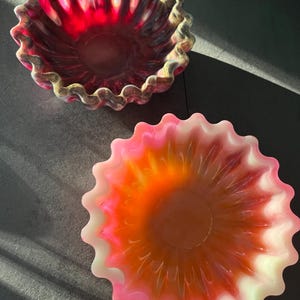 Könnte beinhalten: Zwei dekorative Schalen mit gewelltem Rand. Eine ist tiefrot mit goldenen und braunen Akzenten, die andere ist ein Farbverlauf aus Rosa, Orange und Weiß. Die Schalen sind wahrscheinlich aus Harz oder Glas und stehen auf einer dunklen Oberfläche.