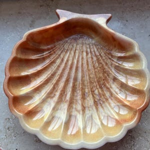 Könnte beinhalten: Eine dekorative Muschelschale mit glänzendem Finish. Die Schale hat ein fächerartiges Muster mit Gold-, Braun- und Weißtönen. Die Ränder sind gewellt und die Schale hat einen kleinen, spitzen Griff. Dieser Artikel eignet sich zum Halten kleiner Gegenstände.