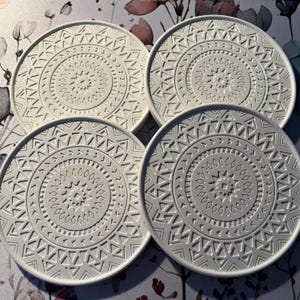 Könnte beinhalten: Vier weiße, kreisförmige Untersetzer mit erhabenem, geometrischem Design. Jeder Untersetzer hat ein zentrales Medaillon, das von konzentrischen Mustern aus Dreiecken und anderen Formen umgeben ist. Die Untersetzer sind quadratisch angeordnet.
