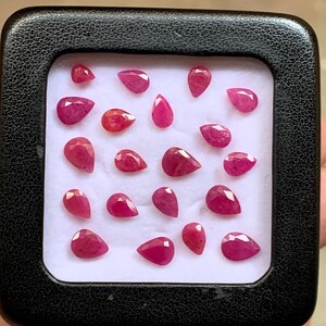 May include: A collection of twenty small, faceted, pear-shaped rubies arranged in a grid pattern on a white foam insert inside a black square display case. Each gemstone exhibits a vibrant, deep pinkish-red hue with visible facets that reflect light.