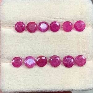 Könnte beinhalten: Zwölf runde, facettierte Rubine, angeordnet in zwei horizontalen Reihen zu je sechs Stück. Jeder Edelstein zeigt einen leuchtenden, tief rosa-roten Farbton mit Brillantschliff, der das Licht auf seinen vielen polierten Oberflächen reflektiert, auf einem cremefarbenen Polster.