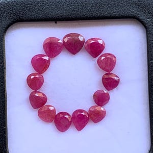 May include: A collection of twelve loose, faceted, pear-shaped rubies arranged in a circular pattern. Each gemstone displays a deep, vibrant magenta-red hue with multiple triangular facets that reflect light across the surface of the stones.