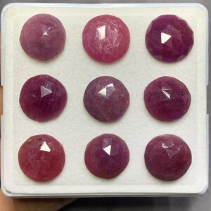 May include: Nine round, faceted rubies arranged in a three-by-three grid. Each gemstone displays a deep pinkish-red hue with a rose-cut surface featuring multiple triangular facets that reflect light, presented in a white square display case.