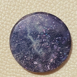 May include: A round, polished gemstone with a deep purple hue, speckled with iridescent flecks of pink, blue, and green. The stone has a textured surface with subtle veining, creating a galaxy-like appearance. The gemstone is set against a neutral background.