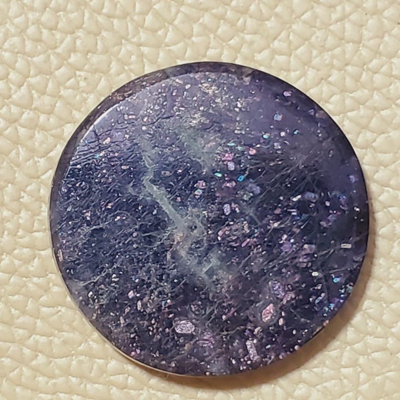 May include: A round, polished gemstone with a deep purple hue, speckled with iridescent flecks of pink, blue, and green. The stone has a textured surface with subtle veining, creating a galaxy-like appearance. The gemstone is set against a neutral background.