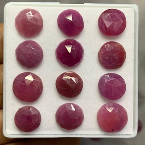 May include: A collection of twelve round, faceted pink and red gemstones arranged in a grid pattern within a white display tray. Each stone features a polished surface with multiple triangular facets that reflect light, showcasing varying shades of ruby.