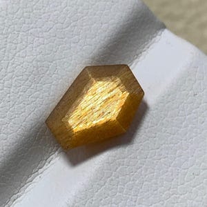 Könnte beinhalten: Ein facettierter, goldbrauner Edelstein mit rechteckiger Form und schimmerndem, reflektierendem Inneren. Der Edelstein hat ein poliertes Aussehen und ist vor einem weißen Hintergrund platziert. Der Edelstein wird wahrscheinlich für Schmuck verwendet.