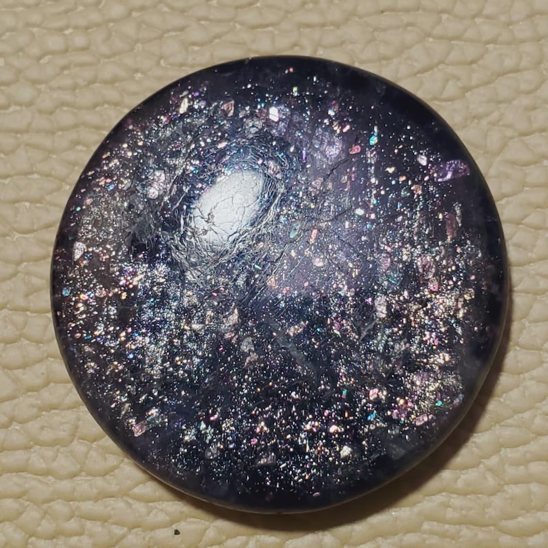May include: A round, dark purple glass cabochon with a galaxy-like appearance. The surface is speckled with iridescent flecks of silver, pink, and blue, creating a shimmering effect. The center has a lighter, almost white, area.