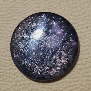 May include: A round, dark blue button with a speckled, iridescent surface. The button has a slightly domed shape and reflects light with a mix of colors, including silver, purple, and green.