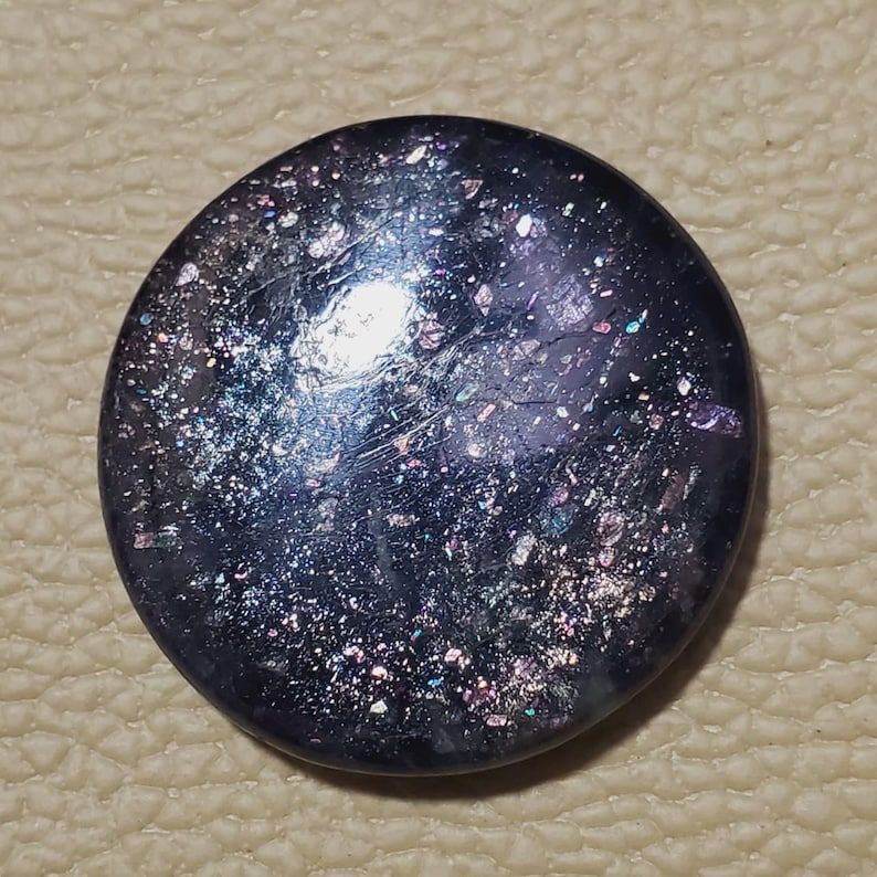 May include: A round, dark blue button with a speckled, iridescent surface. The button has a slightly domed shape and reflects light with a mix of colors, including silver, purple, and green.