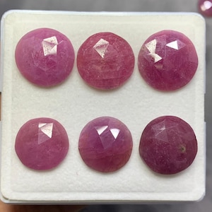May include: Six round, faceted pink rubies arranged in two rows of three on a white display tray. Each gemstone features a circular cut with multiple triangular facets that reflect light, showcasing a deep magenta to pink hue across the stones.