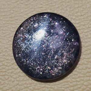 May include: A round, dark blue button with a speckled, iridescent surface. The button has a glossy finish and reflects light with hints of purple, silver, and pink. The button is set against a textured, tan background.