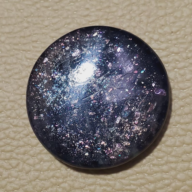May include: A round, dark blue button with a speckled, iridescent surface. The button has a glossy finish and reflects light with hints of purple, silver, and pink. The button is set against a textured, tan background.