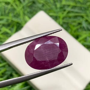 Può includere: Una gemma di rubino ovale e sfaccettata tenuta da una pinzetta. Il rubino è di un colore rosso intenso con molteplici sfaccettature che riflettono la luce. Lo sfondo è verde e bianco sfocato.