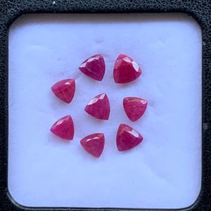 以下が含まれることがあります： 深い赤色をした、三角形にカットされた7つのルース（裸石）のルビー。宝石は白いディスプレイ表面に円形に配置されています。それぞれのルビーには光を反射する複数のファセット（切子面）が施されており、鮮やかで飽和した赤色を際立たせています。